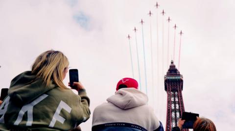 Ground view of an arrowhead formation of nine Red Arrows planes streaming blue, white and red plumes as they fly over Blackpool Tower while, at the bottom of the image, three people take pictures of it on their mobile phones.