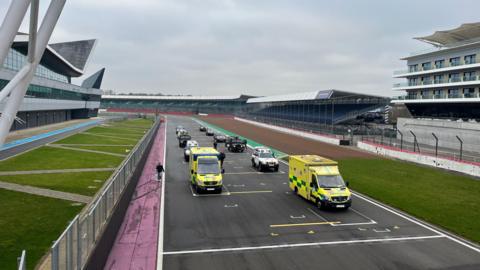 Twelve vehicles, including two ambulances, in formation on a Grand Prix race track.