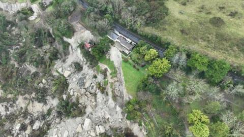 Large rocks and boulders strewn down cliff side buildings and road among trees 