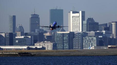 A plane takes off from Logan International Airport in Boston. Skyscrapers and a river are pictured behind the plane.