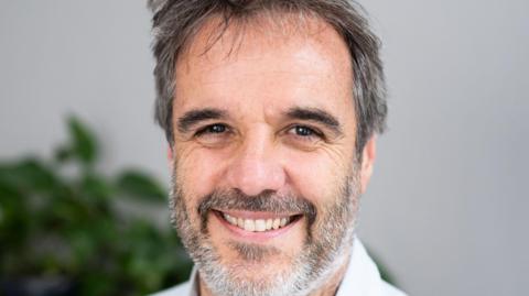 Matthew Taylor smiling with short hair, the background is blurred but there appears to be a green plant behind him.