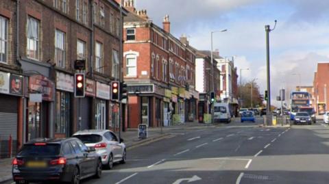 A main road with terraced shops on the left hand side a a large modern supermarket to the right. There are two cars waiting at a set of traffic lights and a bus and other traffic on the right hand side.