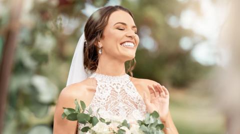 Excited bride in wedding with bouquet in nature park with green trees, bokeh and summer sunshine. Happiness, commitment and dream of a beauty woman with flowers for marriage in outdoor lens flare.