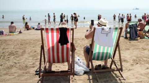 Two people sitting on striped sun loungers on a busy beach on a warm day. Other beach users are wandering around, some are paddling.