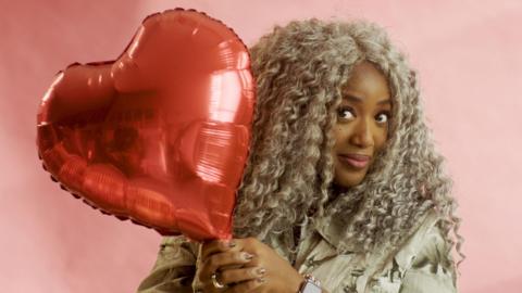 A woman is looking to camera, holding a heart-shaped balloon.