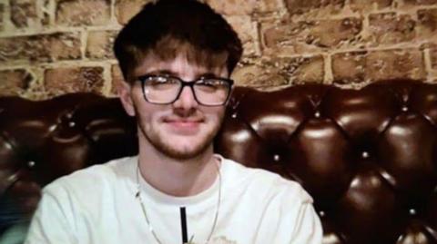 William Webb has dark brown hair and beard and wears a white T-shirt. He sits on a fake leather sofa in front of a brick wall in a bar.