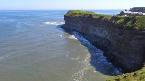 Near vertical cliffs of Jurassic age rocks at Cowbar. The calls of Kittiwakes fill the air here as they find the ledges on this shaded cliff ideal for nesting. To the right are the terraced cottages of Cowbar, separated from the cliff edge by a road, a strip of vegetation and not much else.