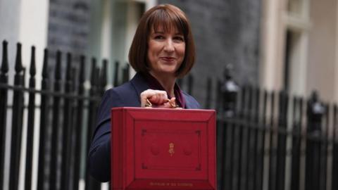 Rachel Reeves in a blue jacket holding the Chancellor's red box ahead of her first Budget. She is outside 11 Downing Street, with the buildings and black iron railings behind her.