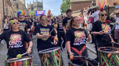 A march through one of Durham's cobbled streets during the Pride celebrations. Women wearing shirts with the words "disco queen" and "disco fever" are beating colourful drums. The people in the crowd wear rainbow-coloured attire, including rainbow afro-style wigs. Some don colourful face paint. In the background, a trade union's banner can be seen waving, and in the distance is Durham Cathedral.