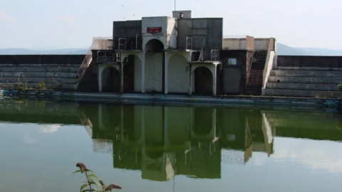 An image of an old art deco Lido building sitting across the murky green water of the lido pool. The building has rows of step seating leading from either side. The water has plants growing in it.