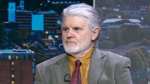 Andrew Boswell, a man with grey white hair. He is sat on the BBC Look East set and is looking away from the camera. He is wearing a mustard coloured shirt, a red and blue patterned tie and grey jacket. 