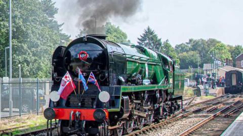 A steam train, painted in green, black and red, with the flags of Poland, the UK and the RAF, pulls out of Tunbridge Wells station.