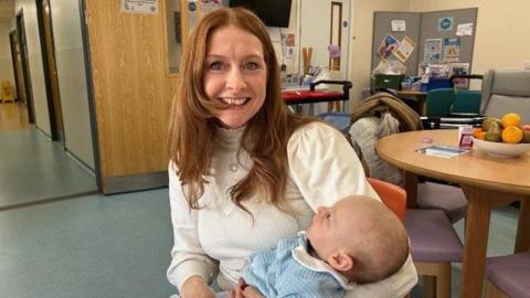 A woman with red hair and a white top holding a baby in blue clothing.