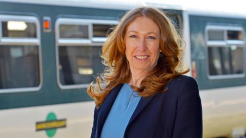 A woman with ginger hair smiling at the camera. She is wearing a light blue shirt and a navy blazer. She is standing in front of a Southern Railway train.