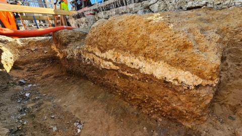 A view from below showing several layers of the roman road underneath the modern tarmac surface of Old Kent Road. Orange tubes are visible to the left of the cross-section