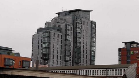 A modern-looking grey and black tower block, significantly taller than any of the other buildings around it, against a white cloudy sky.