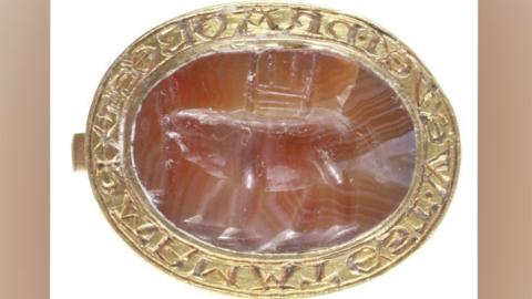 An oval medieval seal matrix on its side, made from gold and a semi-precious gemstone.  Its thin gold rim is etched with carved letters and surrounds the oval pink-toned gemstone, which is carved with an elephant with a castle or howdah on its back. 