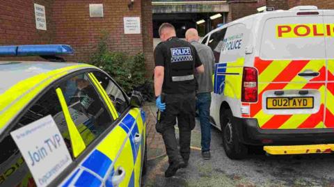 A police officer, between two police vehicles, with his back to the camera leading a man in handcuffs towards a building. The police office has blue gloves on, a black top and black trousers. 