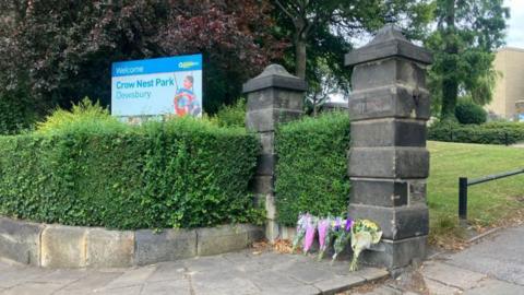 A park entrance with stone pillars and flowers laid out in tribute. A blue sign reads Crow Nest Park.