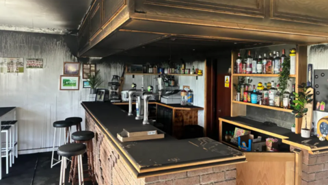 The interior of a clubhouse bar covered in black soot. There is a bar and several stools. 