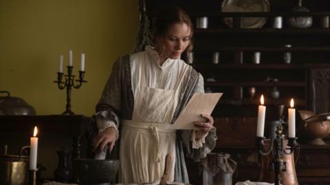 An actress playing Jane Austen. They are in a room lit by candle light and reading from a piece of paper.