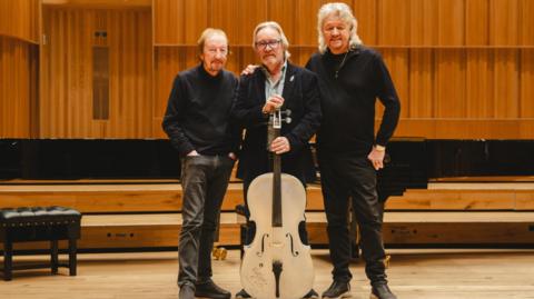 A group of men stand in a auditorium, holding a large silver cello which rests on the floor of the room.