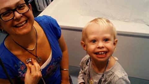 Bea - a young girl with blonde hair - smile. She has a stethoscope around her neck which her mother - a woman with glasses and a blue T-shirt - holds to her chest. The pair smile at the camera.