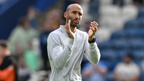 lee grant tilts his head slightly to his left with his brow furrowed in a concentrated way and claps in front of his face. he wears a grey long sleeve polo neck top