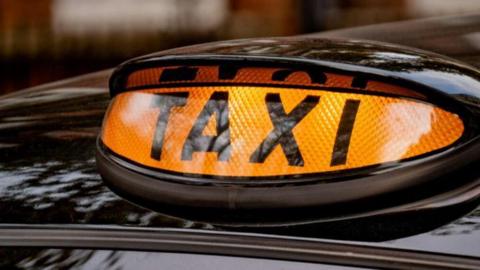 An illuminated orange sign of a taxi. The sign is oval-shaped and attached to the roof of a black car. 