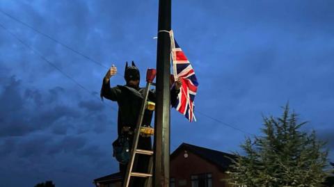 Man in black Batman suit gives a thumbs-up as he ties a Union flag to a street post on a cloudy evening.