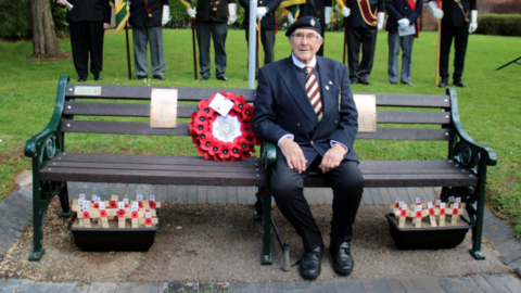 A man wearing clothes including a jacket, a tie and trousers is sitting on a bench next to a wreath and looking at the camera. Eight people are standing up on grass behind him.