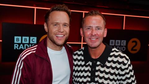Olly Murs is standing on the left wearing a white T-shirt and red jacket with stripes down the arms. He has short brown hair and is smiling at the camera. On the right is Scott Mills on the right wearing a black and white zigzag T-shirt and short brown hair, and is smiling towards the camera. In the back is a sign that says BBC Radio 2.  