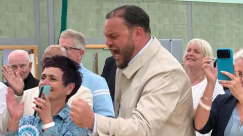 Sam Journet, a Reform councillor wearing a cream jacket, appears to shout and clench his fist in a sports hall, surrounded by other people clapping.