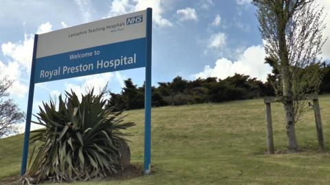 A sign saying 'Welcome to the Royal Preston Hospital' and 'Lancashire Teaching Hospitals NHS Trust' is on a steep bank of grass, with a plant beneath it and trees nearby.