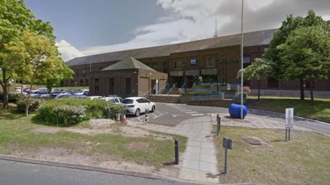 Peterborough's Thorpe Wood Police Station - a two-story brick building with an access road, grass verges and trees in front of it.