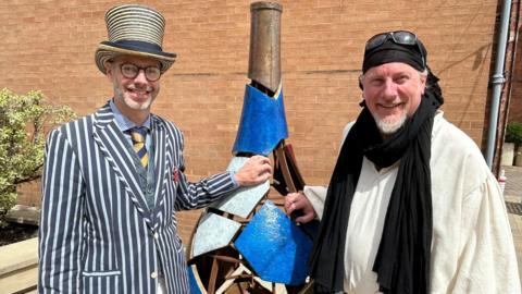A man with glasses and a short grey beard is wearing a yellow- and black-striped top hat, with a pinstripe blazer over a waistcoat and a blue and yellow tie. On his left is a piece of art which looks like a cracked vase. On the other side of the artwork is a man with a black bandana and black scarf over a white tunic. Both men are resting their hands on the artwork.
