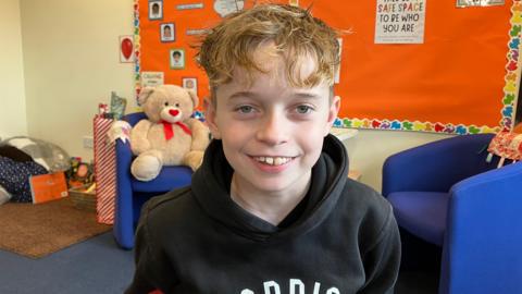 A young boy with blond wavy hair smiles at the camera. He is in a classroom with a bright red display board behind him.