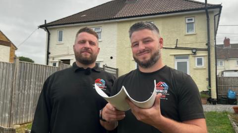 Two young men with dark beards and short cropped dark hair face the camera. They are both wearing black T-shirts with a red, white and blue company logo on them and one is holding a section of white drainpipe. Behind them are two houses, with a light wood garden fence between them