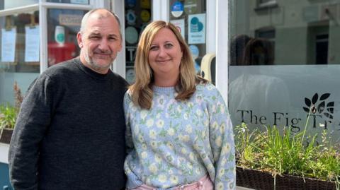 Ryan Marsland and a colleague stood directly outside their shop. They are both looking at the camera and are smiling. There are window boxes with greenery in front of the large glass windows which has the company's name and logo on. 