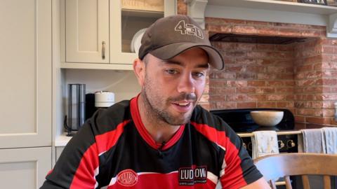 Lewis Ludlow in a Gloucester Rugby red and black top and a brown cap, sitting in a kitchen.
