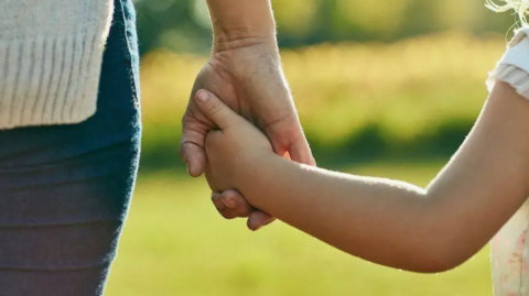A stock image of an an adult and a child holding hands outside. They are standing side by side but we can only see their hands, forearms and a small part of the side of their bodies. 