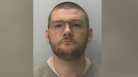 A police mugshot of Ricky Stubberfield. He is wearing a grey jumper and glasses. He has a beard and is staring straight at the camera. 
