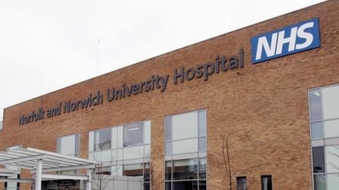 A sign at the front of the hospital which reads, Norfolk and Norwich University Hospital NHS. The hospital has brown bricks and large windows