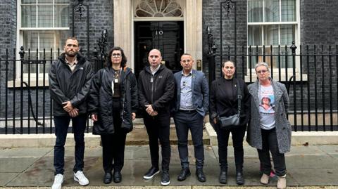 The families of the hostages standing outside the door of Downing Street - there are six people standing in a row with the famous black door behind them. Steve Brisley, Ayelet Svatitzky, and Sharone Lifschitz are all present, with a separate woman wearing a t-shirt with a picture of what is presumably her relative.