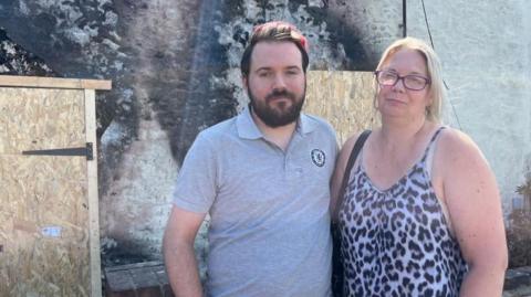 Garry Berger is wearing a grey top with a logo on the left breast. He has dark hair, with red tints and a full dark beard and moustache. His aunt, Christina Steele is wearing a leopard print vest top. She has long blonde hair and is wearing glasses. They are standing outside the house owned by his parents.