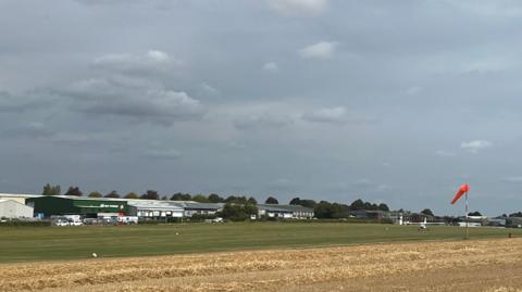 A view of the airfield under a grey sky. A mown yellow crop field with a green airstrip next to it. Aviation buildings in the background. One small plan on the grass airstrip.