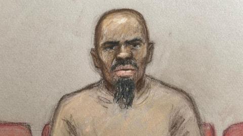 Sketch of Suleiman who looks dead ahead of him and wears a beige jumper. He has a scraggly beard and sits on a row of red seats in front of a blank wall 