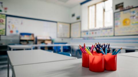 An empty school classroom with tubs of pencils on a desk and an array of seats and tables