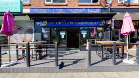 The front of a premises called 'Taylor's Sports Bar and Grill', as indicated by a blue sign above the glass door. Tables and parasols sit on a small decked area outside.
