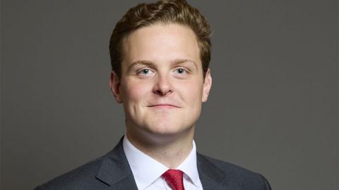 Head and shoulders image of Oliver Ryan standing in front of a dark grey background. He has wavy brown hair and is smiling. He is wearing a dark grey suit, white shirt and red tie